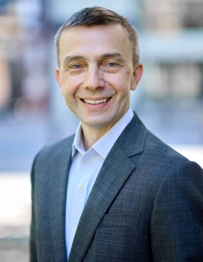 Portrait of a smiling man in a gray blazer and light blue shirt, outdoors with a blurred urban background.