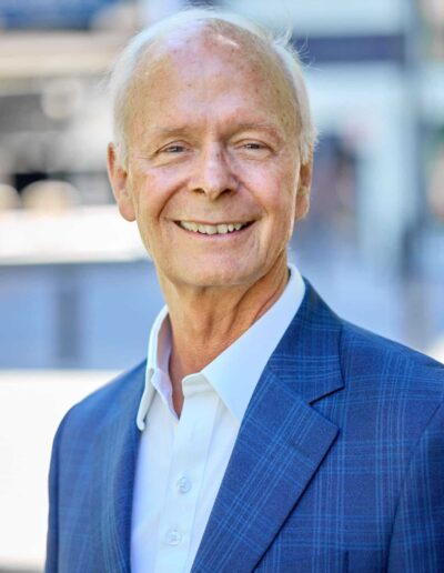 Smiling older man in a blue plaid suit outdoors, blurred urban background.