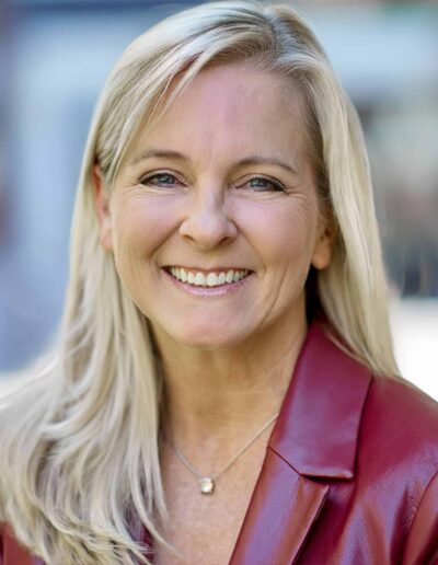 Smiling blonde woman in a burgundy leather jacket outdoors, facing the camera.