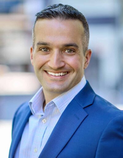 Smiling man in a blue suit outdoors, head and shoulders portrait with blurred background.