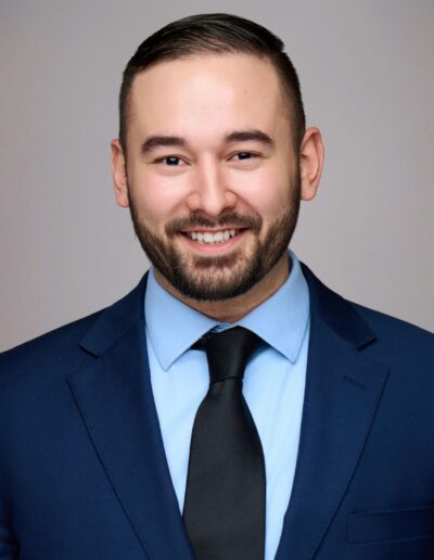 Smiling man in a navy suit, light blue shirt, and black tie, looking at the camera against a neutral background (professional portrait).