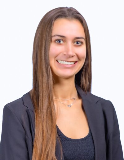 Professional headshot of a smiling woman with long brown hair, wearing a dark blazer and navy top against a light background.
